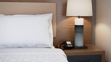 A neatly made bed with a white pillow and a patterned blanket, a bedside table with a lamp, and a small drawer.