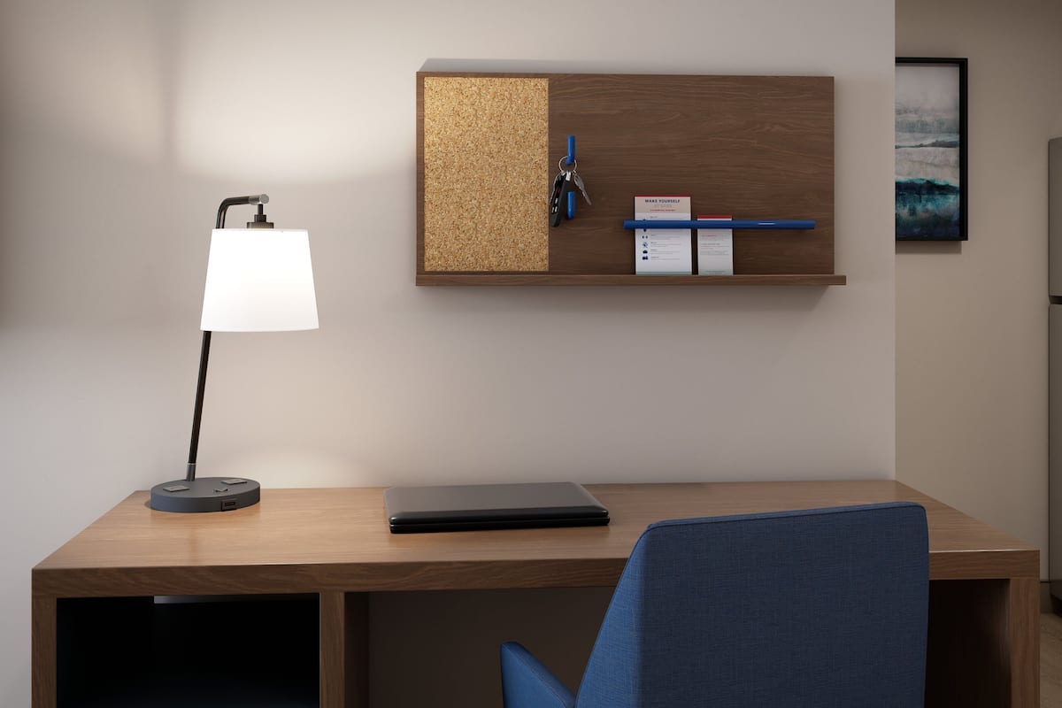 Ein moderner Schreibtisch mit einem Laptop, einer Schreibtischlampe, einem Korkwandorganisator mit Schlüsseln und einem Notizblock sowie einem gerahmten Bild an der Wand.