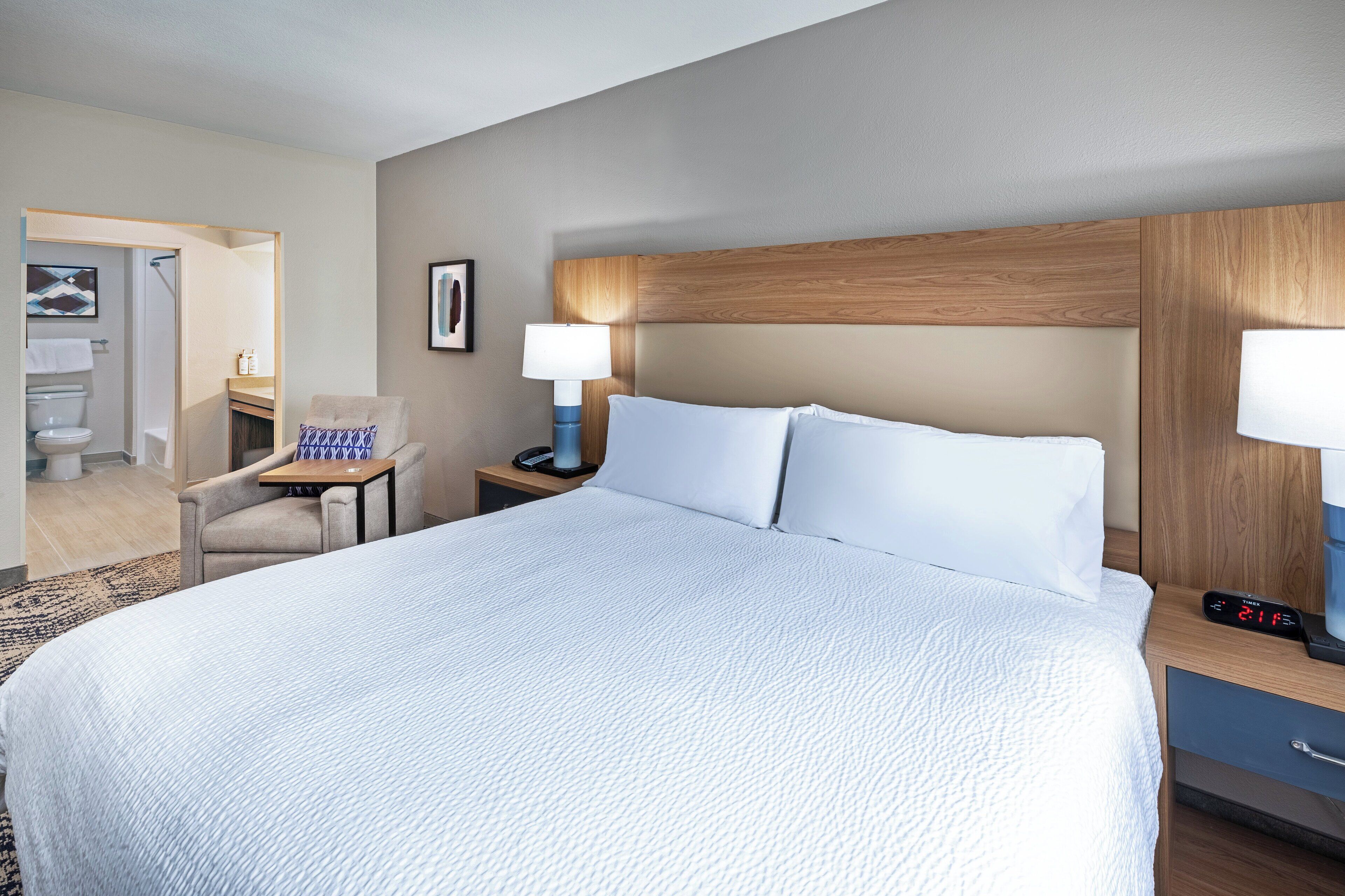Une chambre d’hôtel avec un grand lit, une table de chevet avec une lampe, une chaise et une petite table sur laquelle se trouve une bouteille.