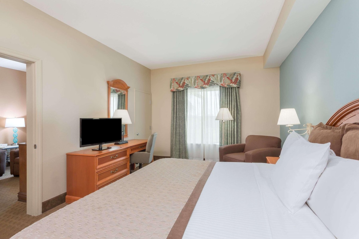 Quarto de hotel com uma cama grande, uma escrivaninha com televisão, uma cadeira e uma janela com cortinas.