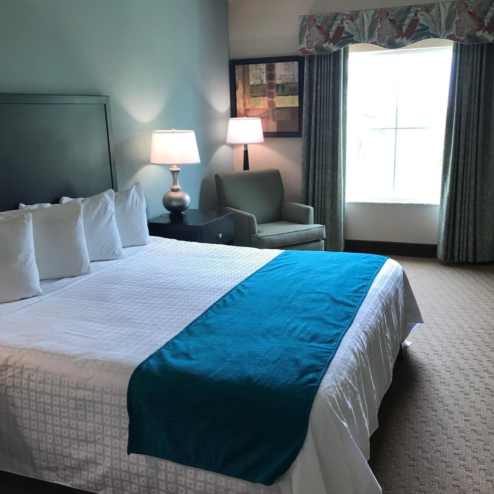 Quarto de hotel com cama grande, uma cadeira, mesa de cabeceira, abajur e janela com cortinas.