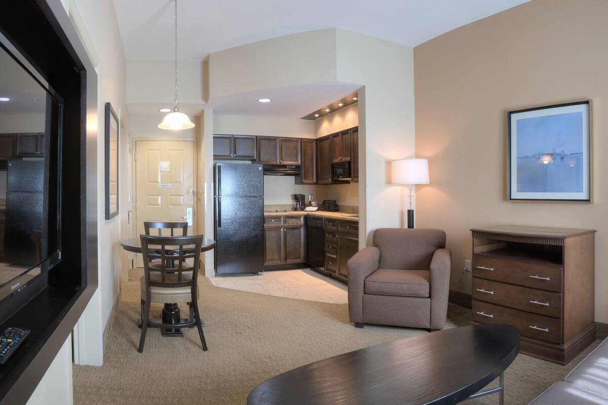 A modern living room with a flat-screen TV, a dining area with a table and chairs, a sofa, and a kitchen in the background.