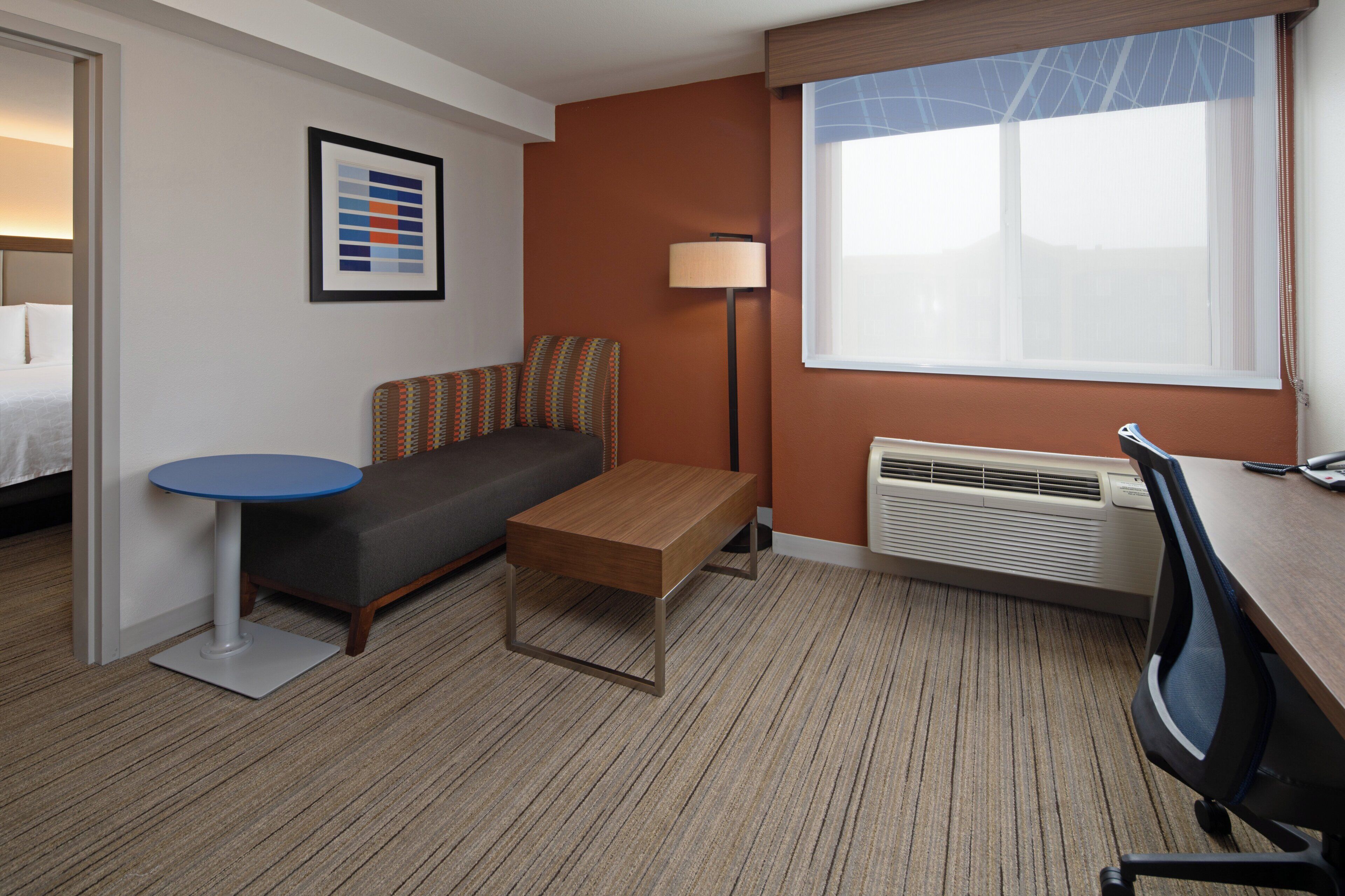 A hotel room with a bed, a sofa, a small table, a desk, a lamp, and a window with blinds.
