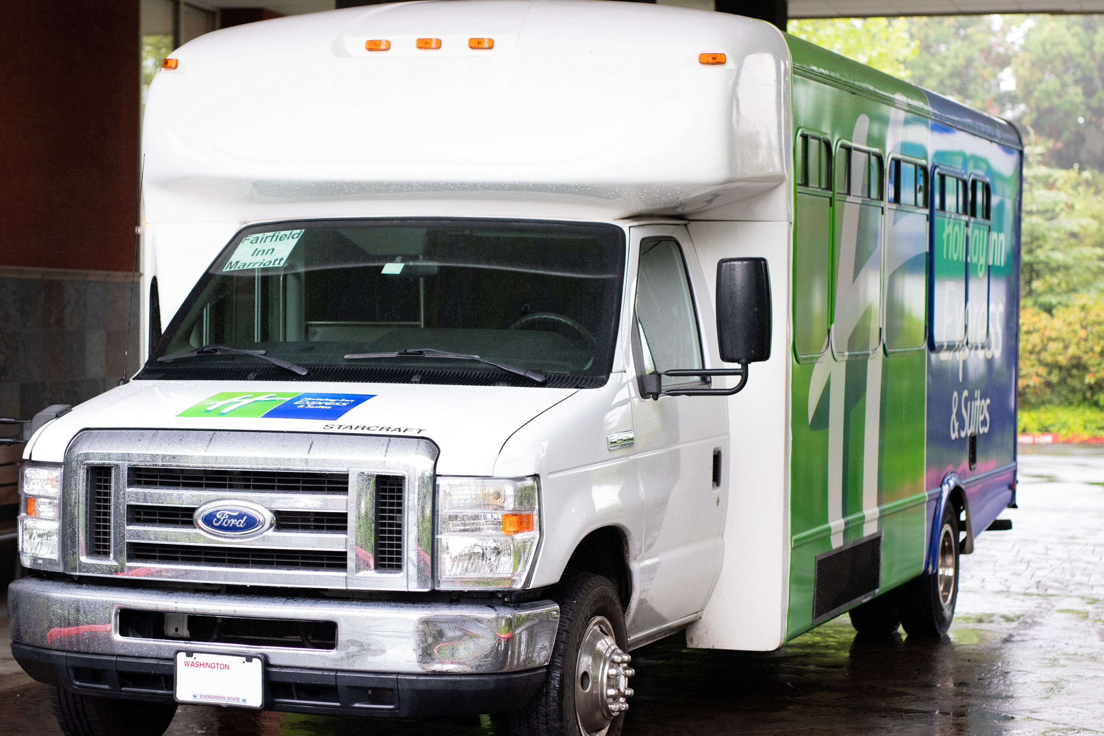 A white Ford transit van with a Fairfield Inn Marriott sign on the windshield, parked under a shelter.