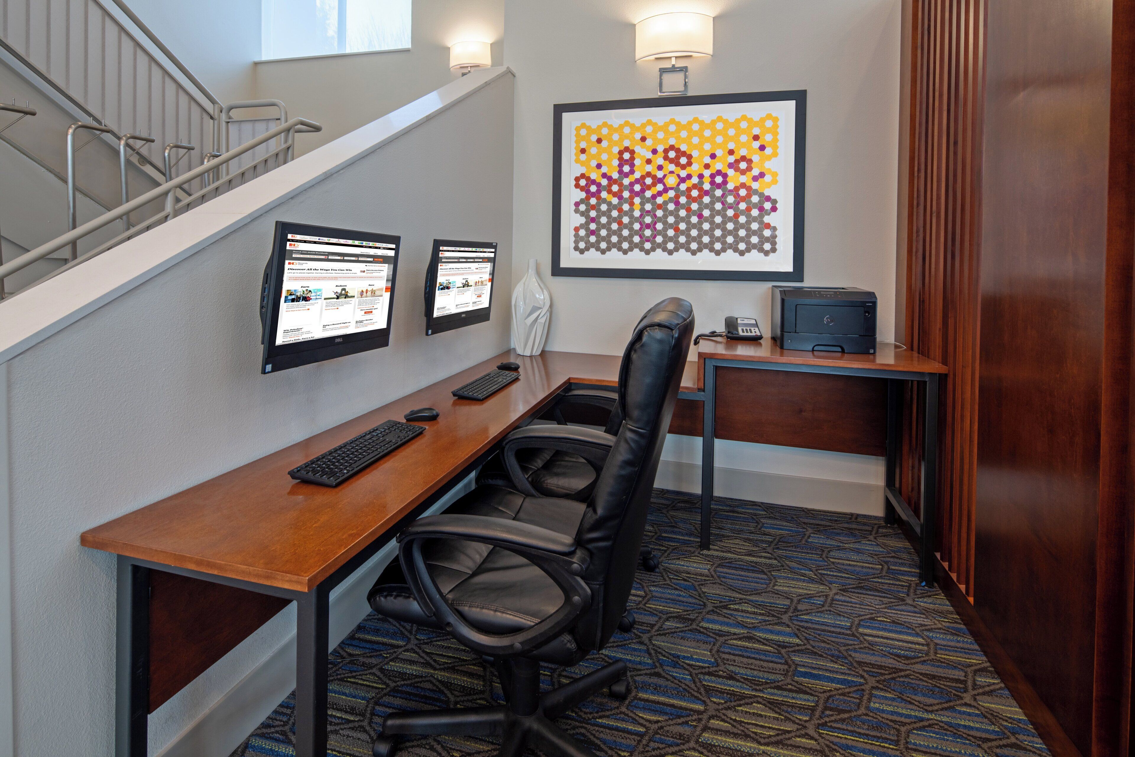 A hotel room with a desk, two monitors, a chair, a telephone, and a framed artwork on the wall.