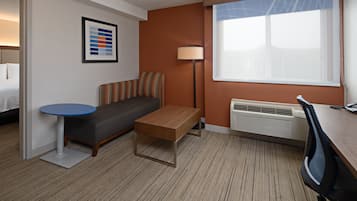 A hotel room with a bed, a sofa, a small table, a desk, a chair, a lamp, and a large window.