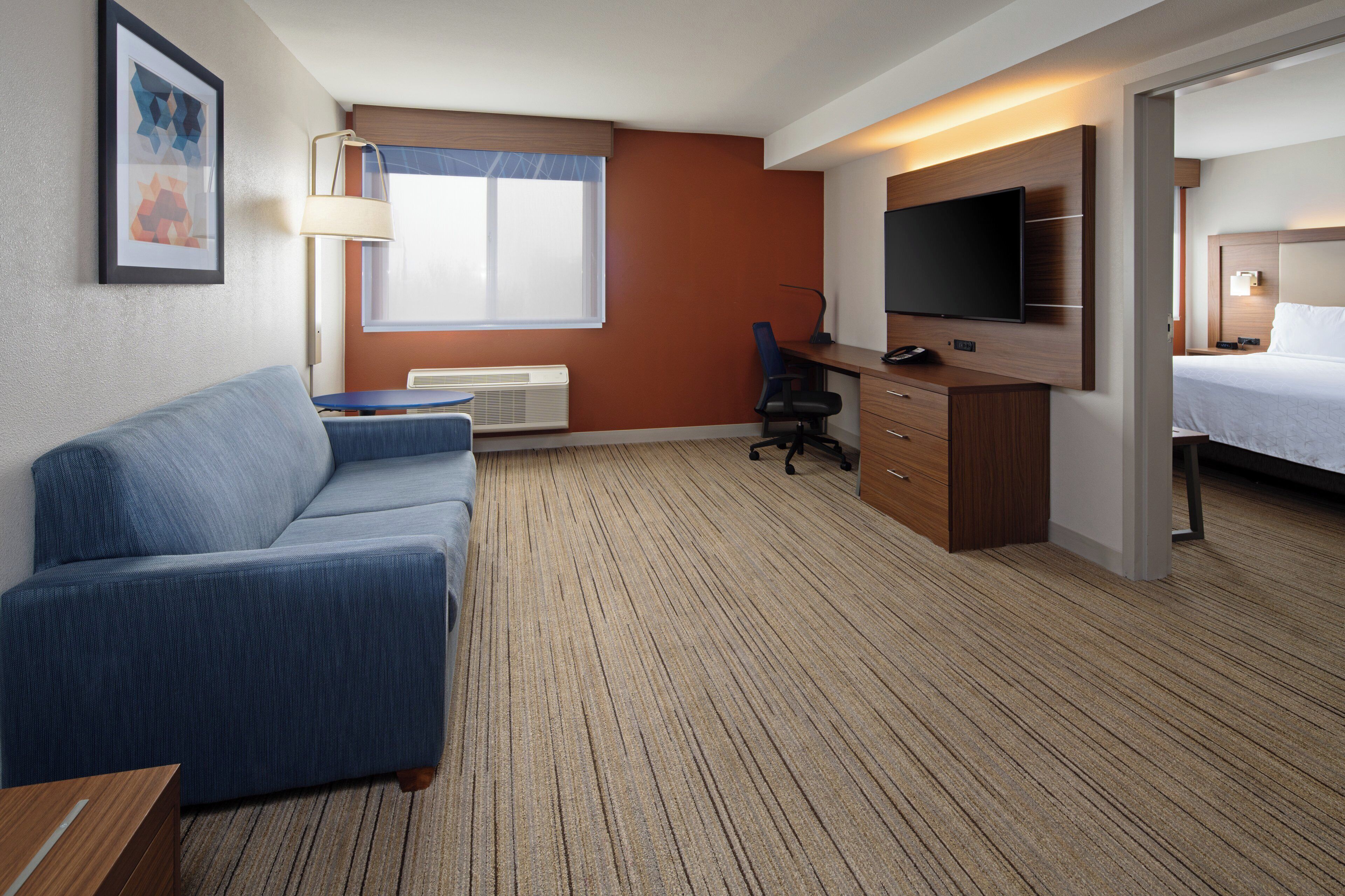 A hotel room with a blue sofa, a desk with a computer, a flat-screen TV, a bed, and a window with blinds.