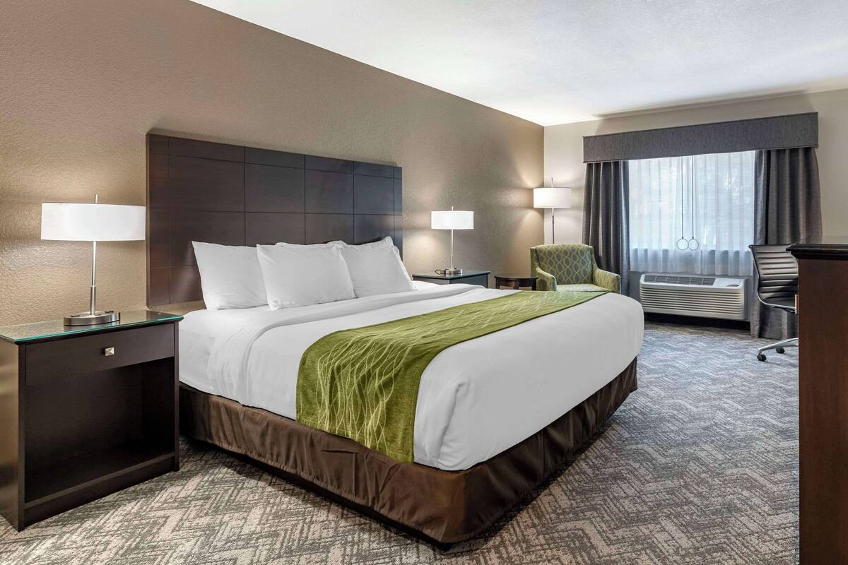 Habitación de hotel con una cama grande, dos mesitas de noche con lámparas, una silla y un ventanal con cortinas.
