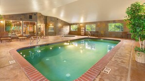 An indoor swimming pool with a brick border, lounge chairs, and a potted plant.