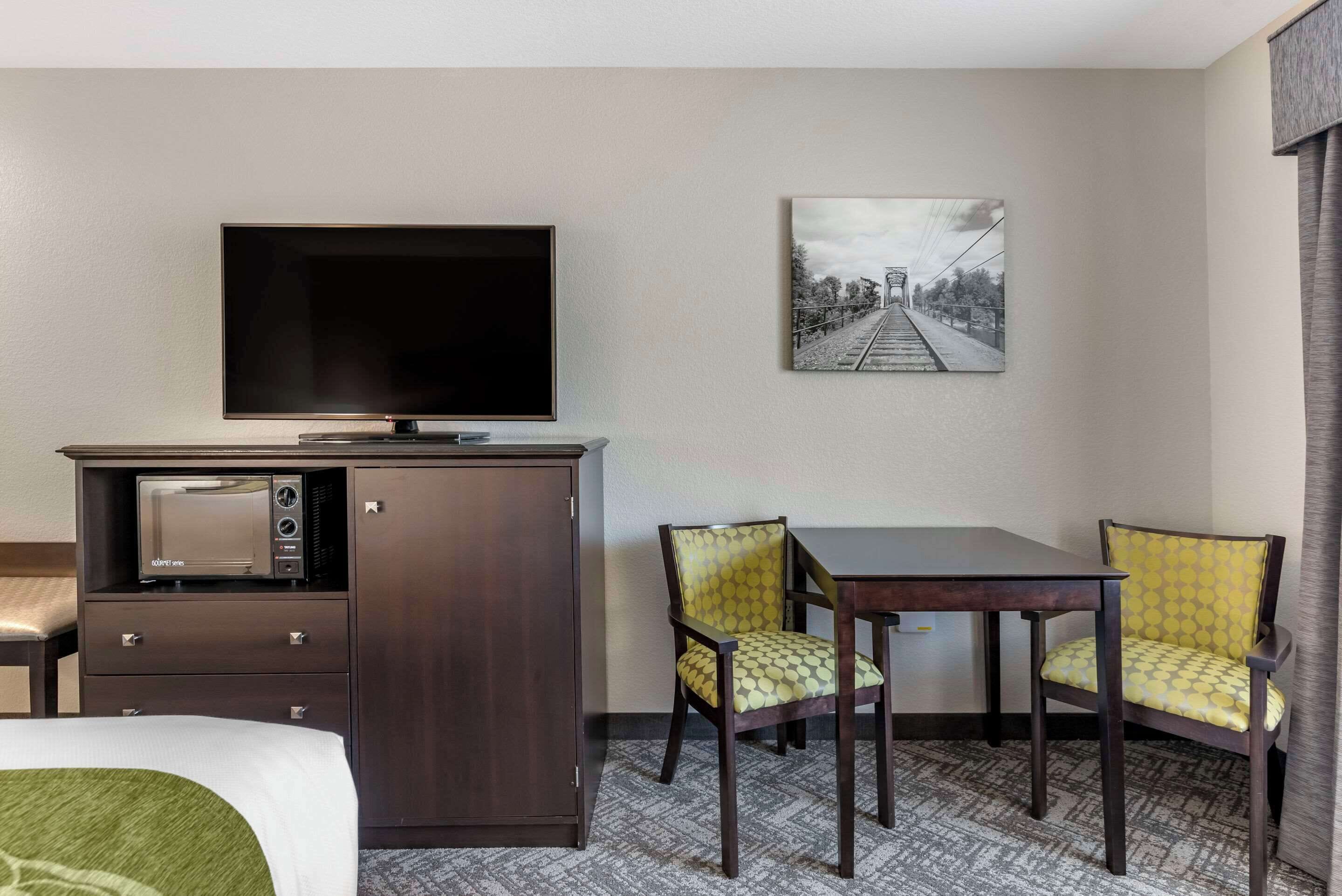 Quarto de hotel com cama, televisão, micro-ondas, pequeno armário, duas cadeiras, mesa e um quadro na parede.