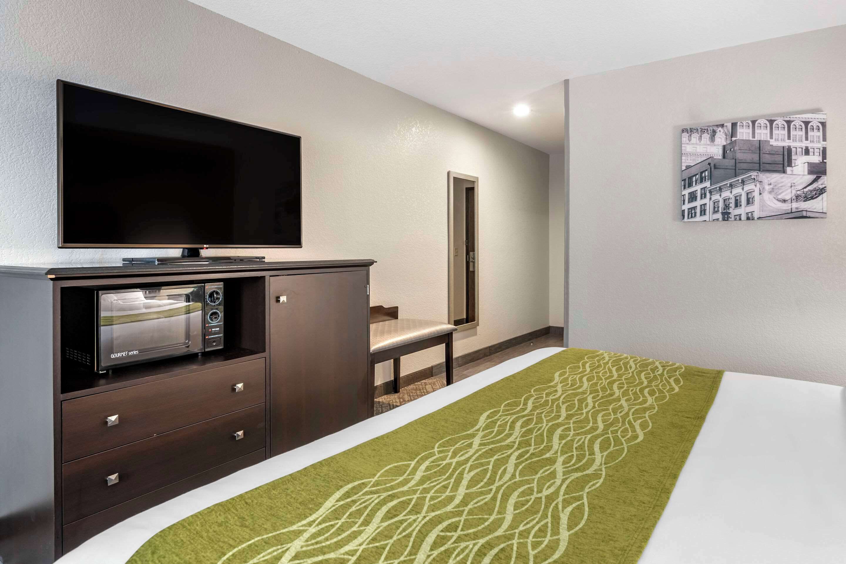 A hotel room with a flat-screen TV, a microwave, a bench, and a patterned rug.