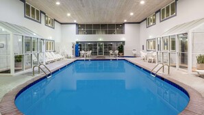 Una piscina interior con paredes de vidrio transparente, sillas de descanso blancas y piso de baldosas.