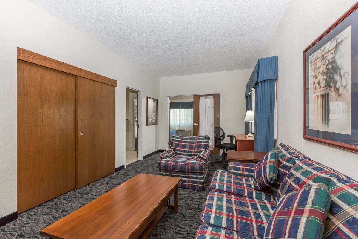 Una habitación de hotel con un conjunto de sofás, una mesa de madera, un escritorio con una silla, un armario y un cuadro en la pared.