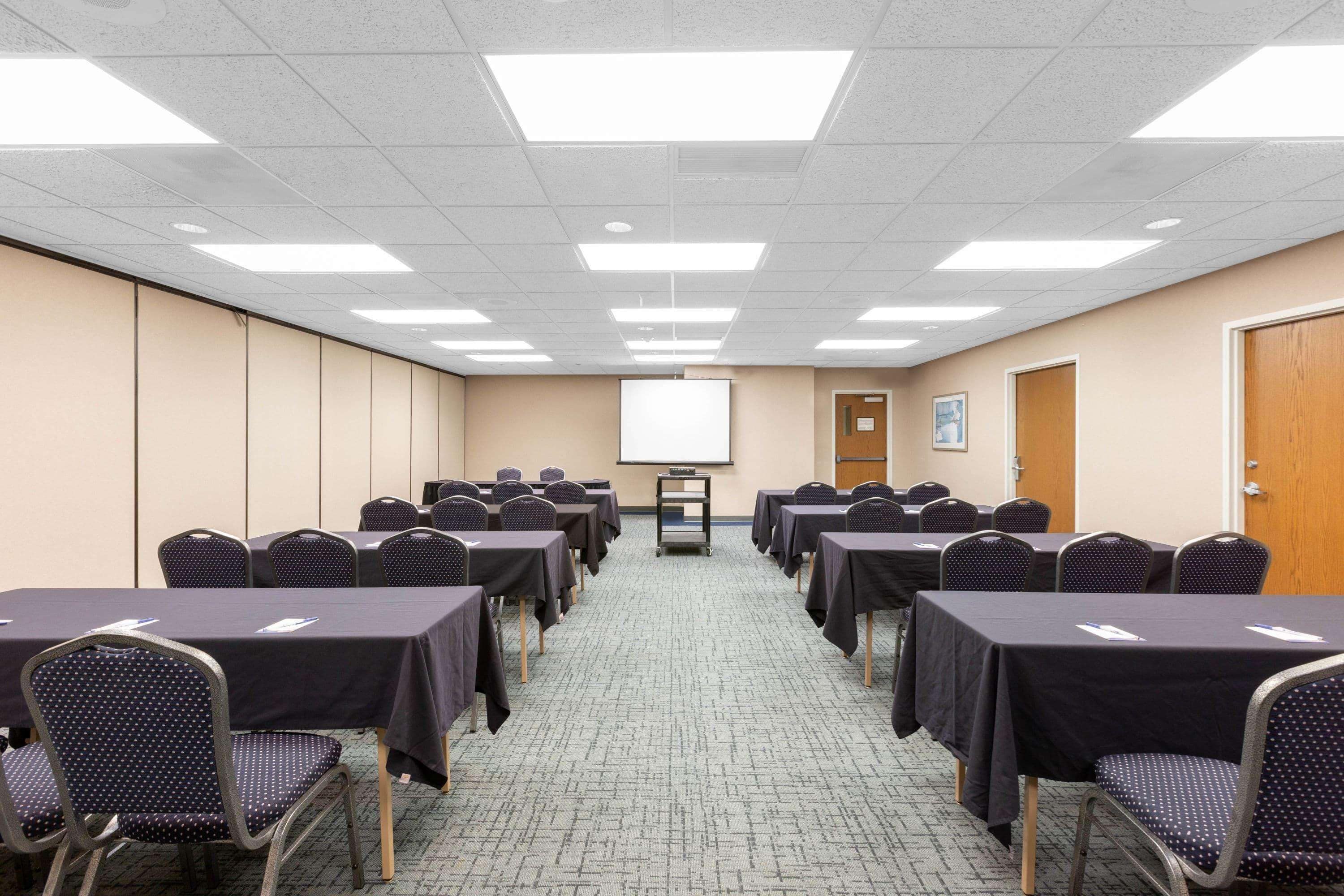 Un salón de conferencias con filas de mesas y sillas, una pantalla de proyector y una puerta a la derecha.