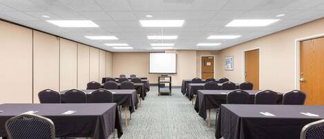 Un salón de conferencias con filas de mesas y sillas, una pantalla de proyector y una puerta a la derecha.