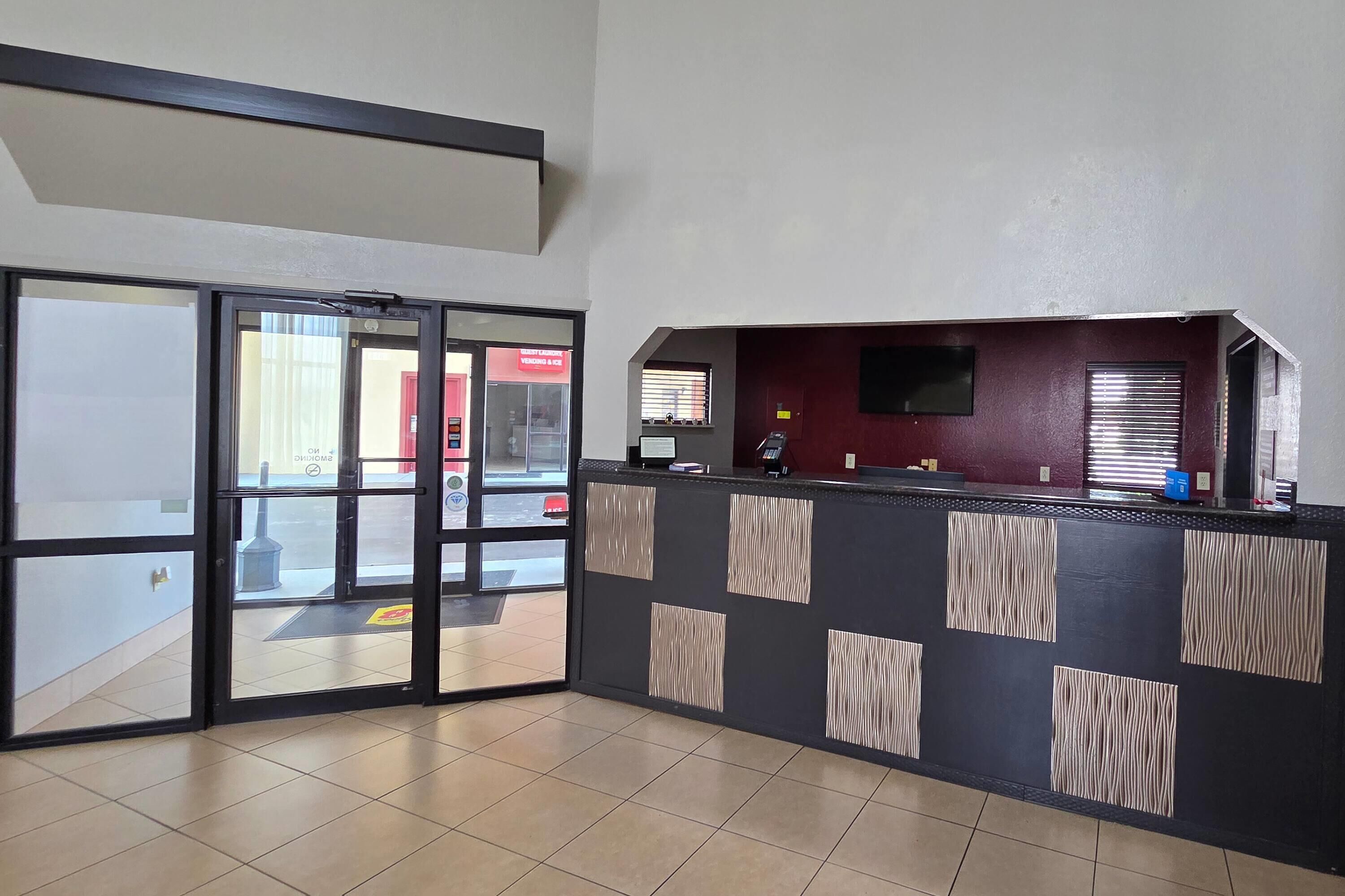 Un área de recepción con entrada de puerta de vidrio, un mostrador con televisor de pantalla plana y suelo de baldosas.
