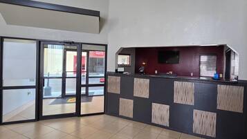 Un área de recepción con entrada de puerta de vidrio, un mostrador con televisor de pantalla plana y suelo de baldosas.