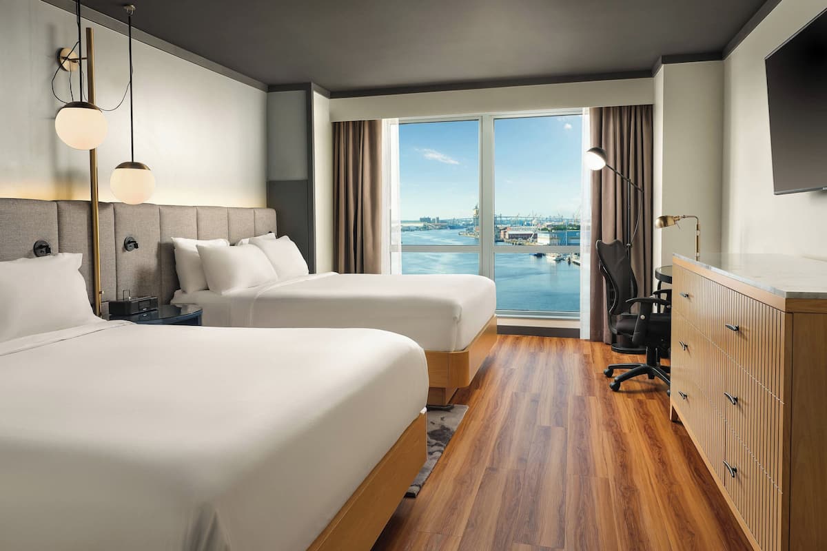 Une chambre d’hôtel avec deux lits, un plancher en bois, un bureau avec une chaise, une télévision et une vue sur l’eau et le paysage urbain.