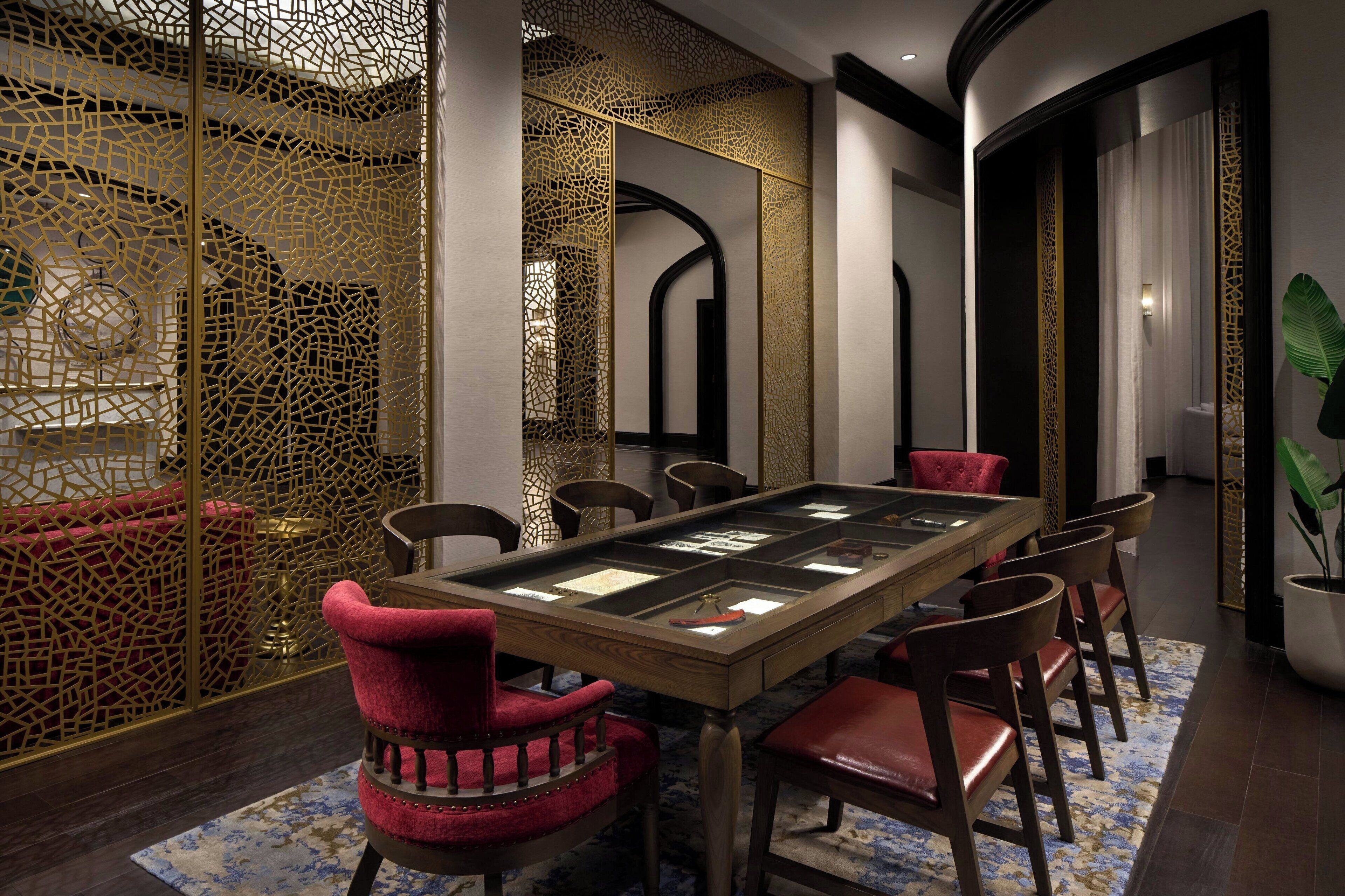 Un espace repas comprenant une longue table, des chaises à revêtement rouge et un paravent décoratif orné d’un motif géométrique.