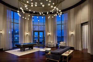 A spacious room with a pool table, comfortable seating, and large windows offering a view of the cityscape.