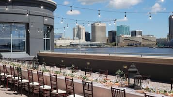 An outdoor dining area with a city skyline view, tables set with white tablecloths and chairs, and string lights overhead.