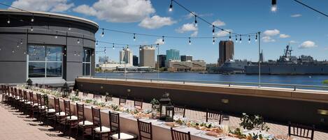 An outdoor dining area with a waterfront view, neatly arranged tables and chairs, and string lights.