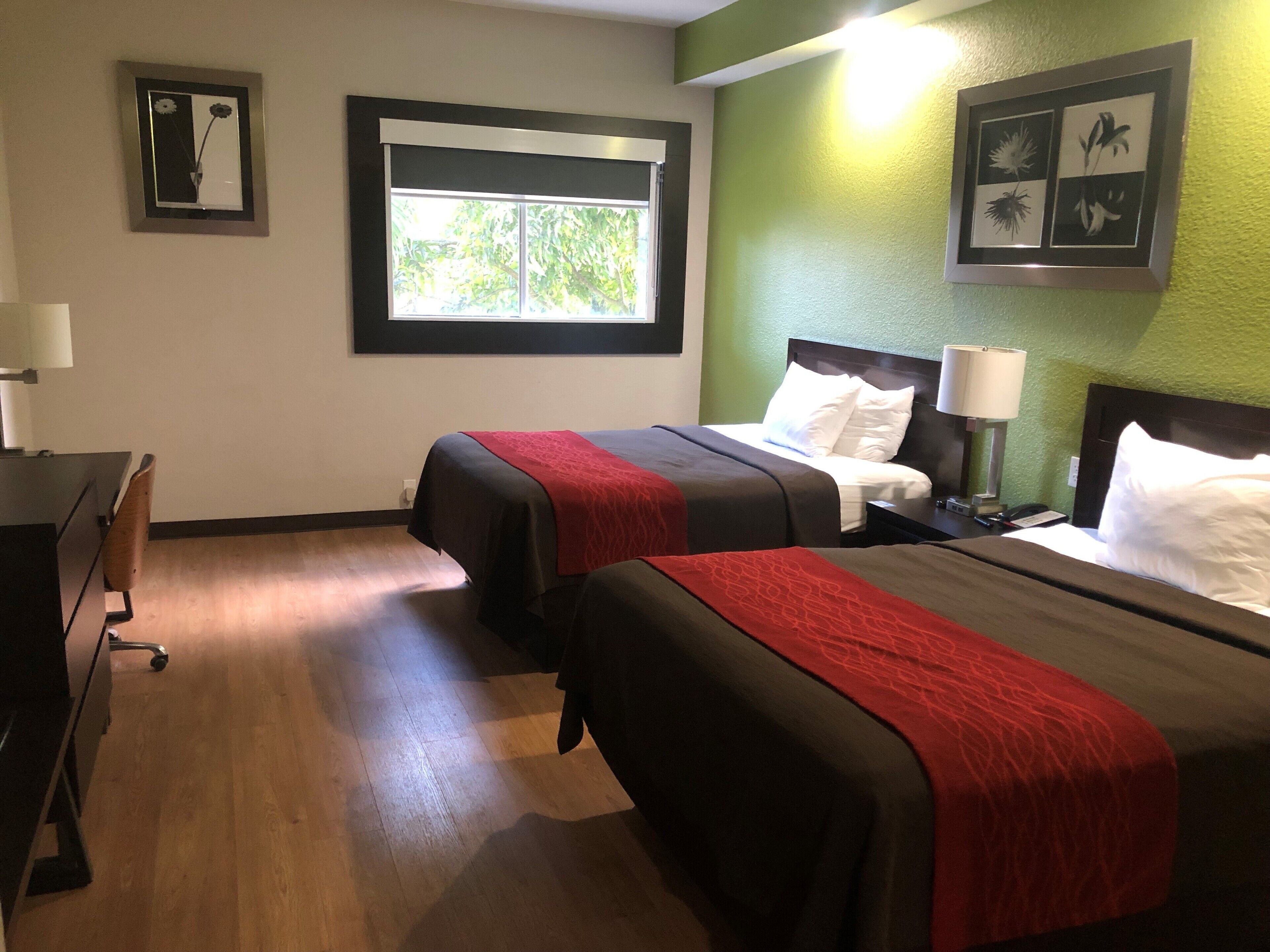 A hotel room with two beds, a desk, a chair, a window, and framed artwork on the walls.