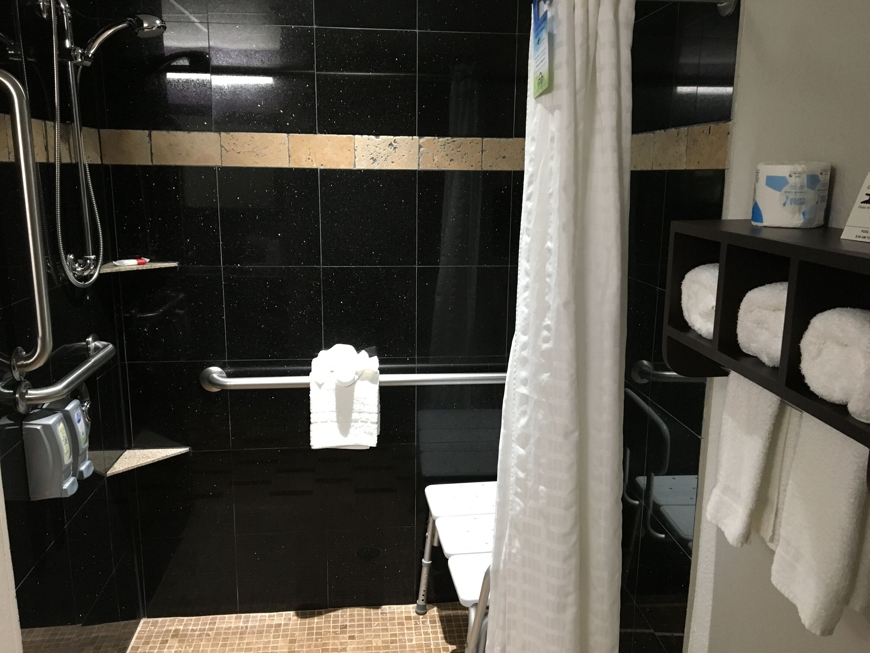 Un baño con paredes de azulejos negros, una ducha con cabezal de ducha de mano, una barra de apoyo, una toalla blanca en un perchero y un estante con toallas blancas.