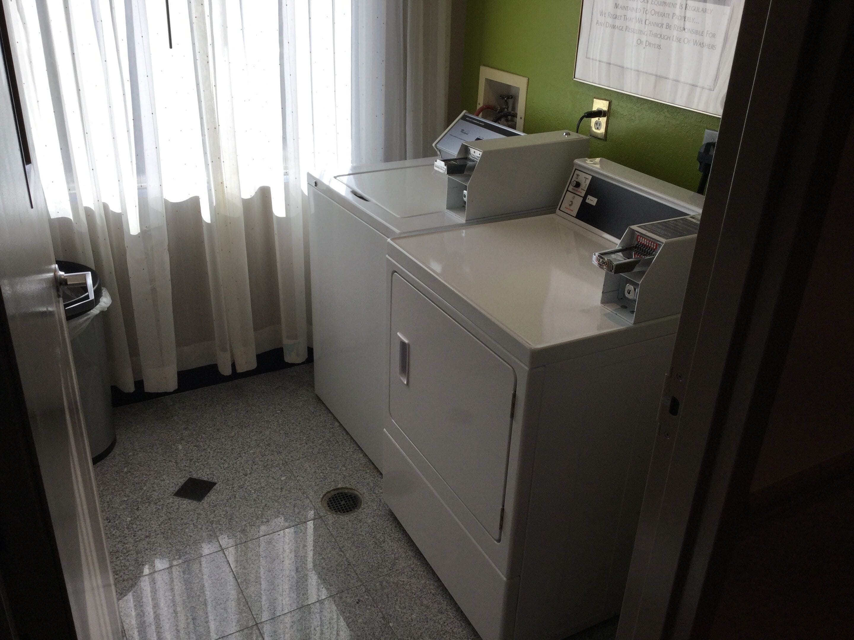 A laundry area with a washing machine and dryer, a small window with curtains, and a green wall with a notice.
