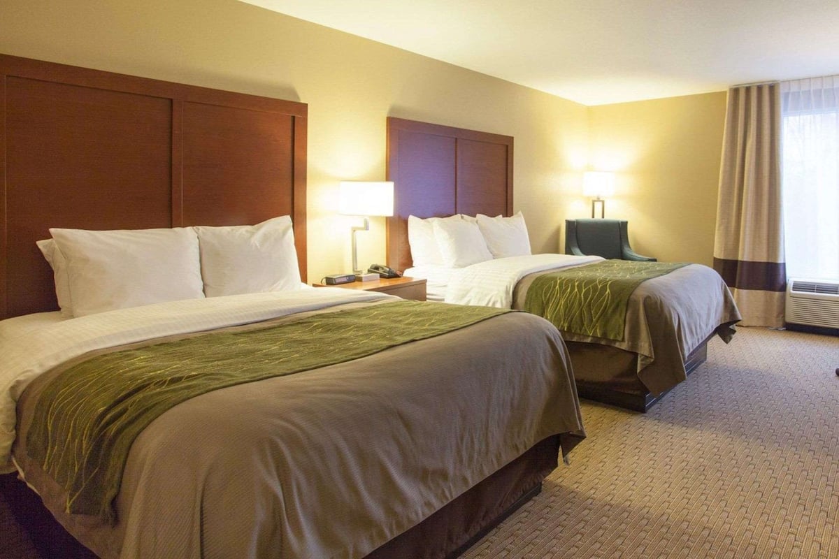 Habitación de hotel con dos camas, una mesita de noche con lámpara, una silla y un ventanal con cortinas.
