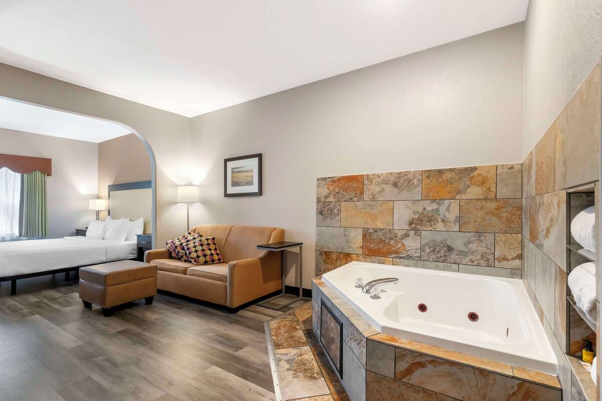 A modern hotel room with a large bed, a sofa, a small table, a desk, and a bathtub.