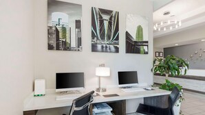 A modern office space with two desks, each with a computer monitor, keyboard, and mouse. There are two chairs, one black and one with wheels. A printer is on the floor, and a lamp is on the desk. The walls are decorated with framed pictures of bridges. - Best Western Plus Des Moines West Inn & Suites (Clive)