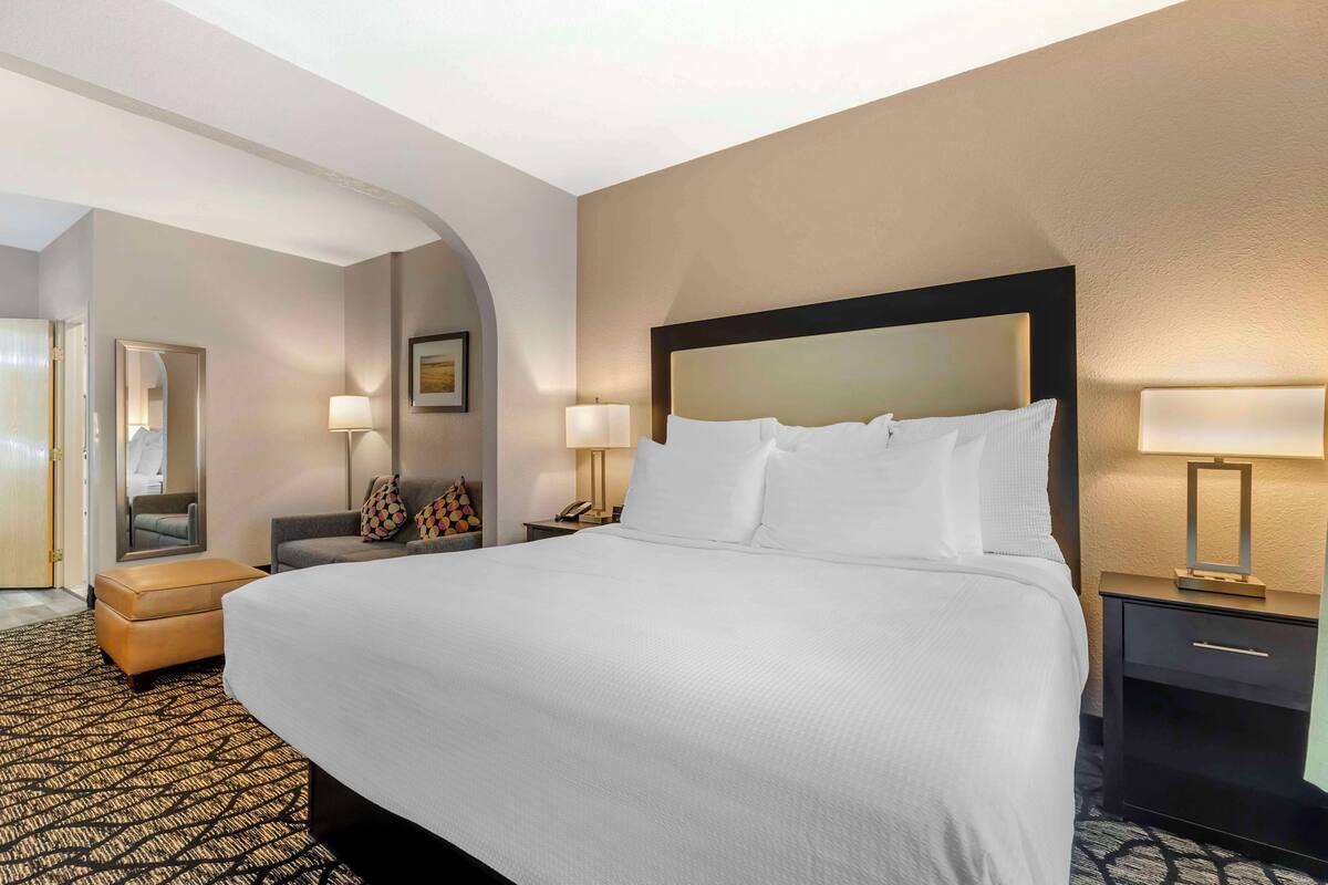 A hotel room with a large bed, a nightstand with a lamp, a small table with a lamp, a sofa, and an arched doorway.