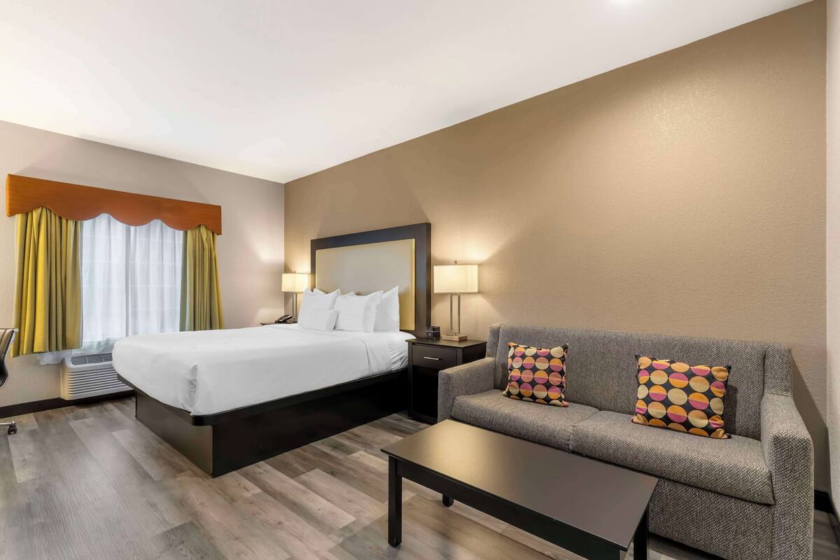 A hotel room with a bed, a sofa, a coffee table, a nightstand, a lamp, and a window with curtains.