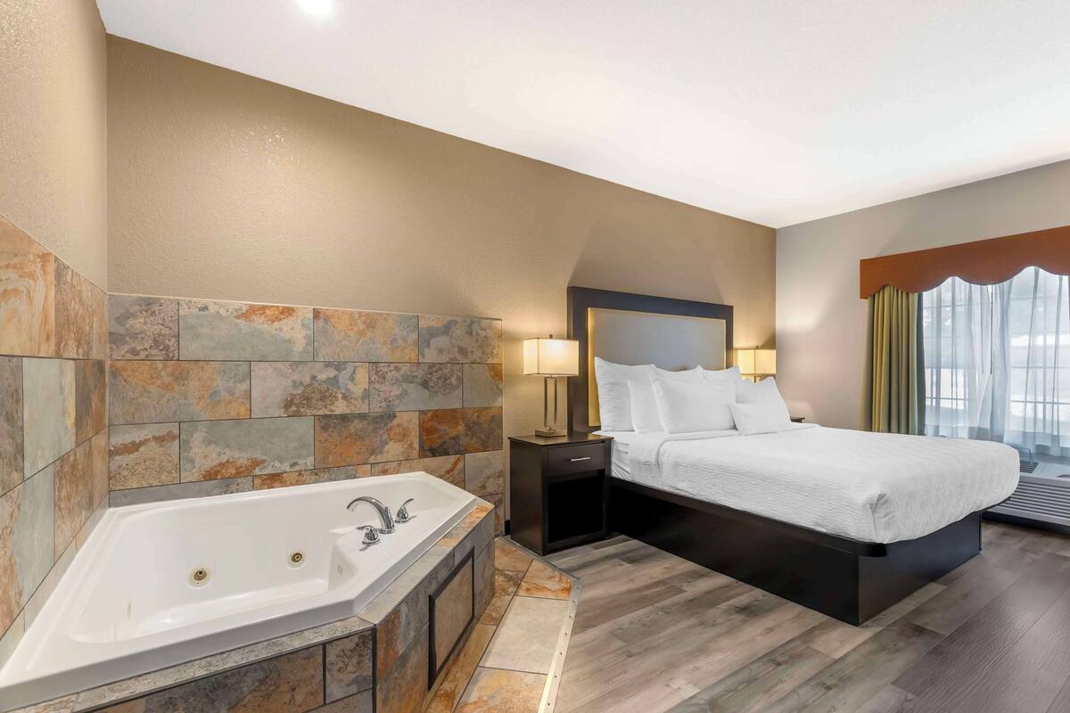 A modern hotel room with a large bathtub, a bed with white linens, and a nightstand with a lamp.