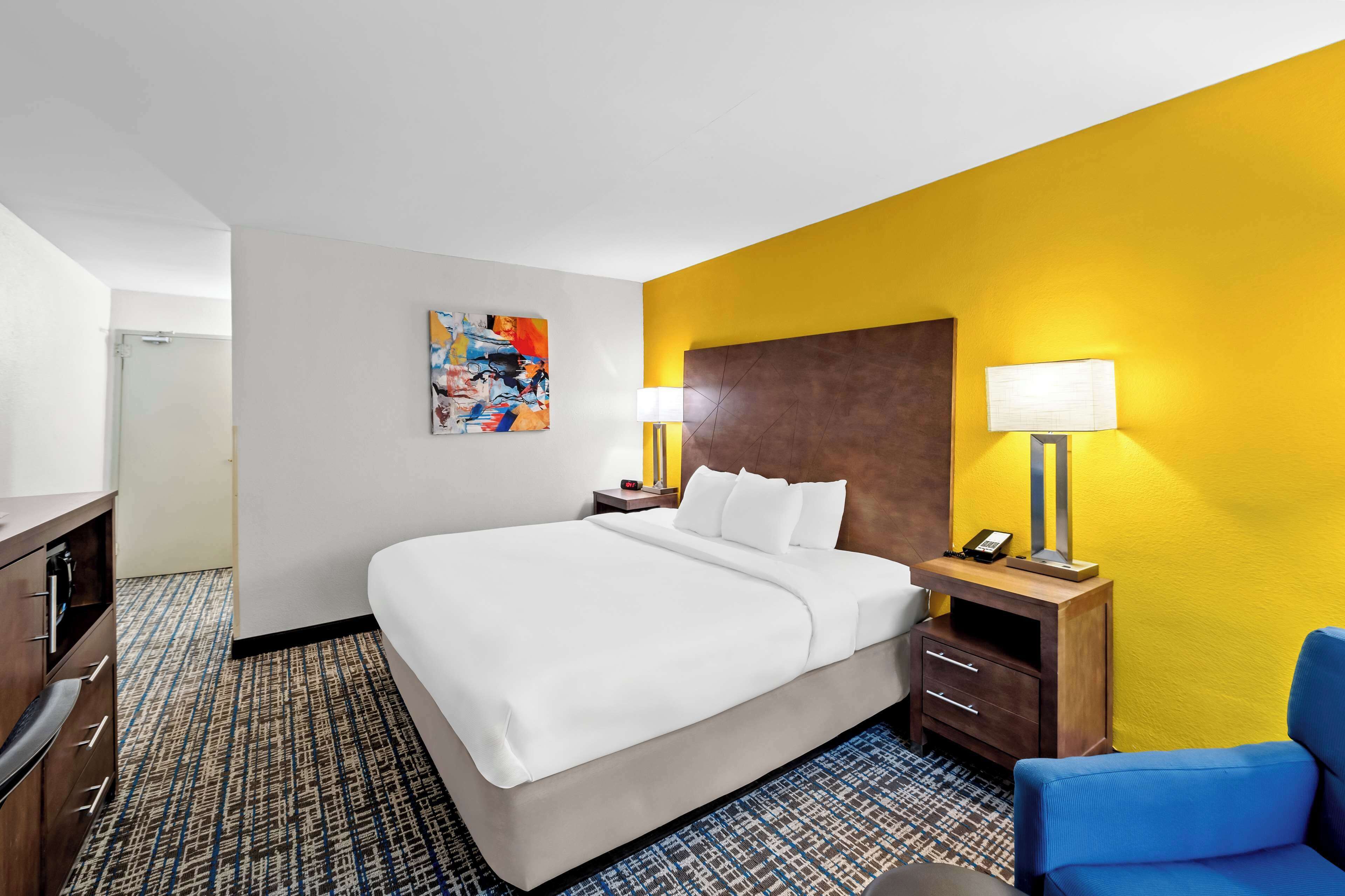 Una habitación de hotel con una cama grande, una mesita de noche con lámpara, una silla azul y un cuadro abstracto de colores en la pared.