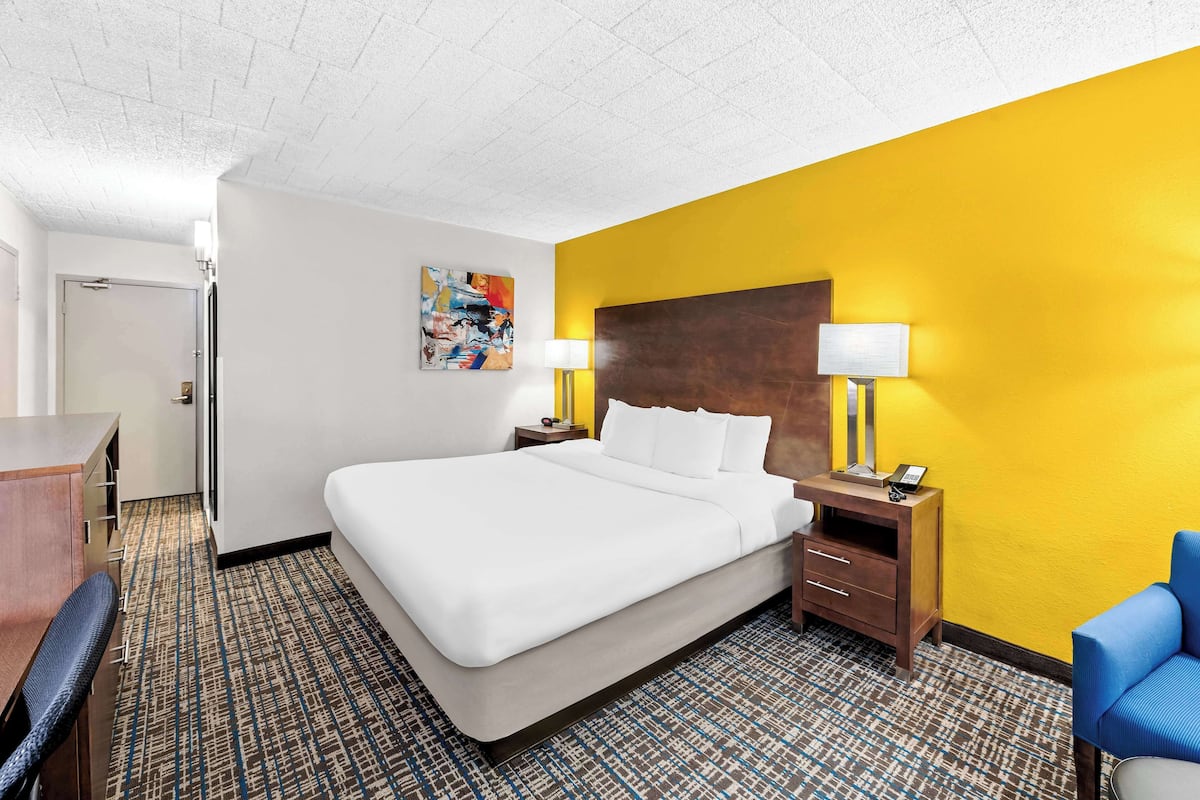 A hotel room with a large bed, a desk, and a chair. There is a painting on the wall and a lamp on the nightstand.