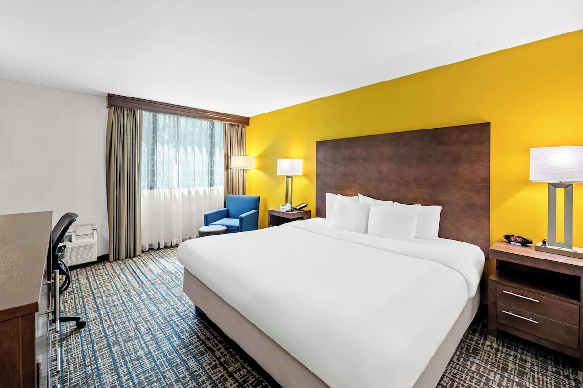 A hotel room with a large bed, a desk with a chair, a desk lamp, and a window with curtains.