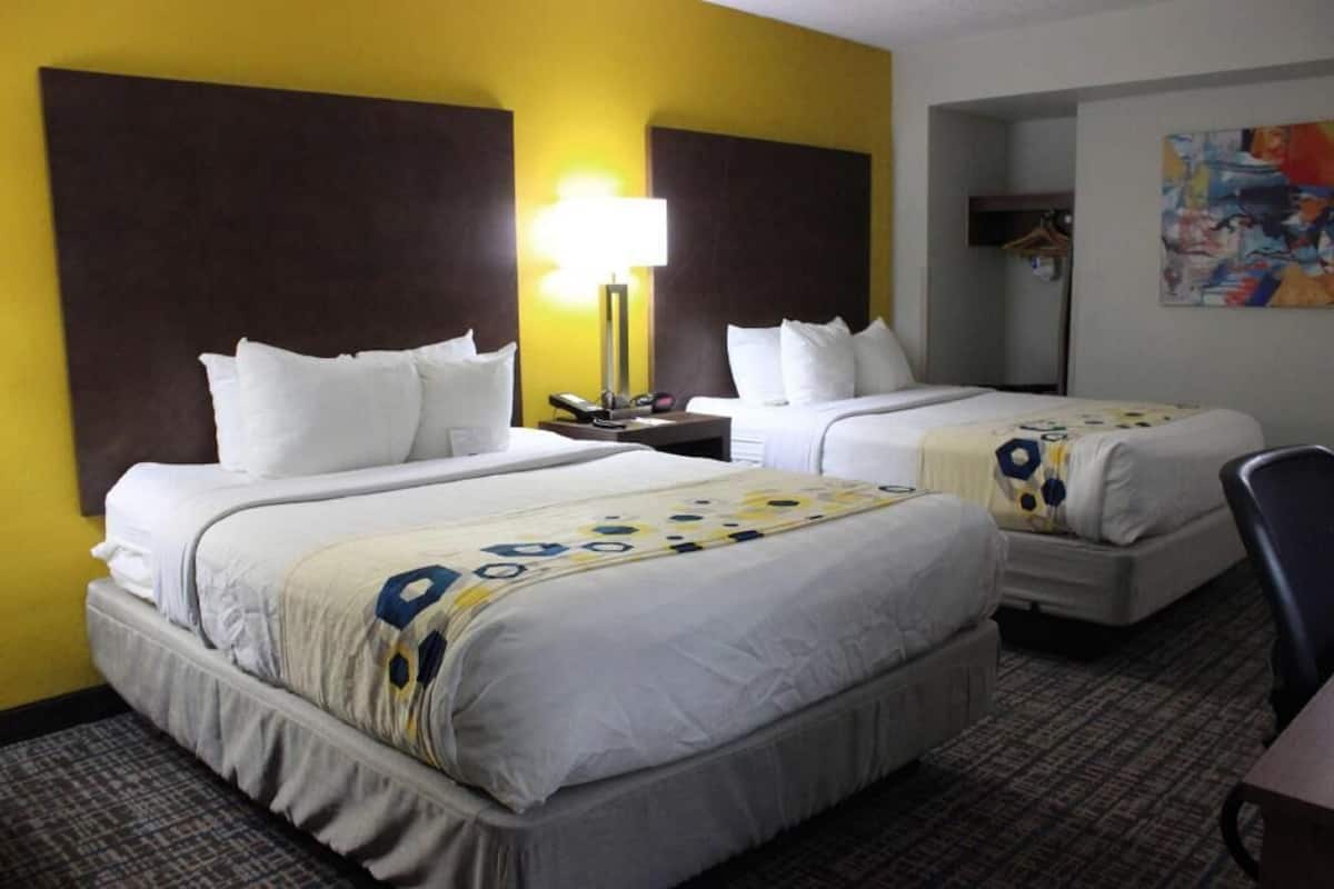 A hotel room with two beds, a nightstand, a lamp, and a painting on the wall.