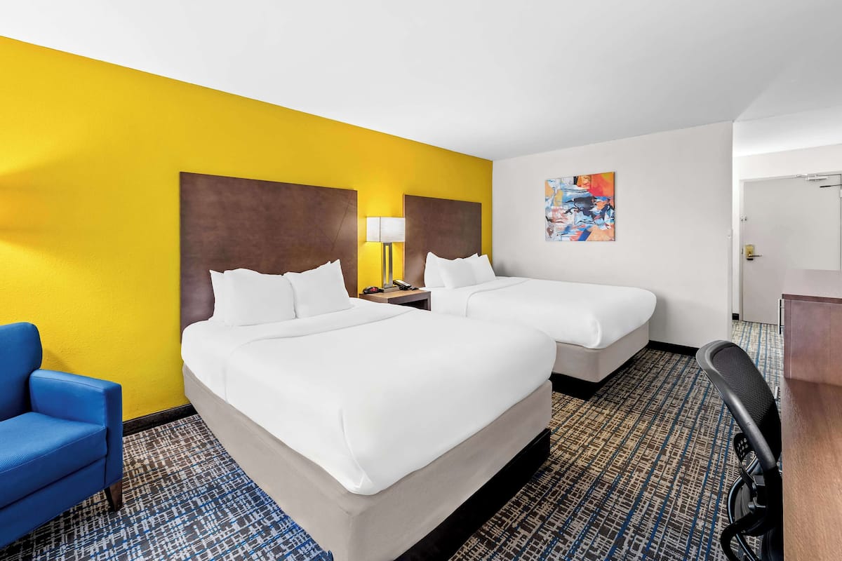 A hotel room with two beds, a blue armchair, a desk, and a patterned carpet.