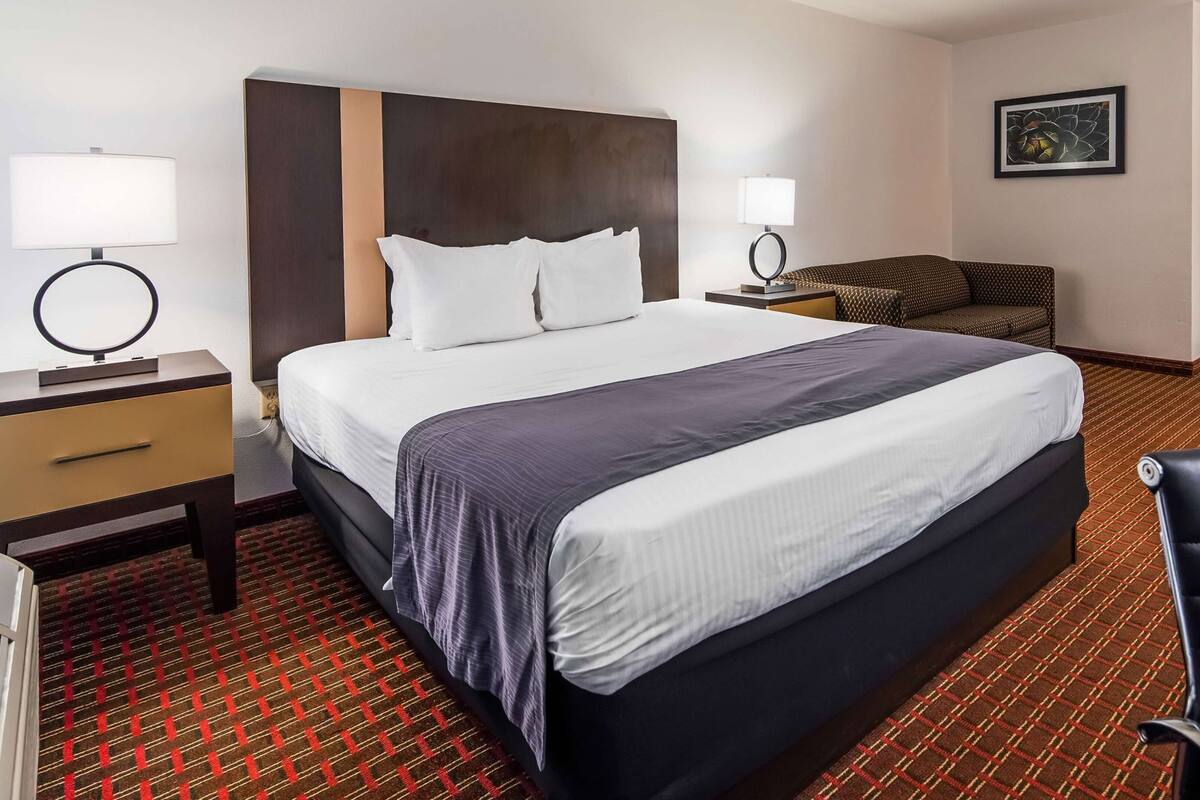 A hotel room with a large bed, a desk with a lamp, a chair, and a small sofa.