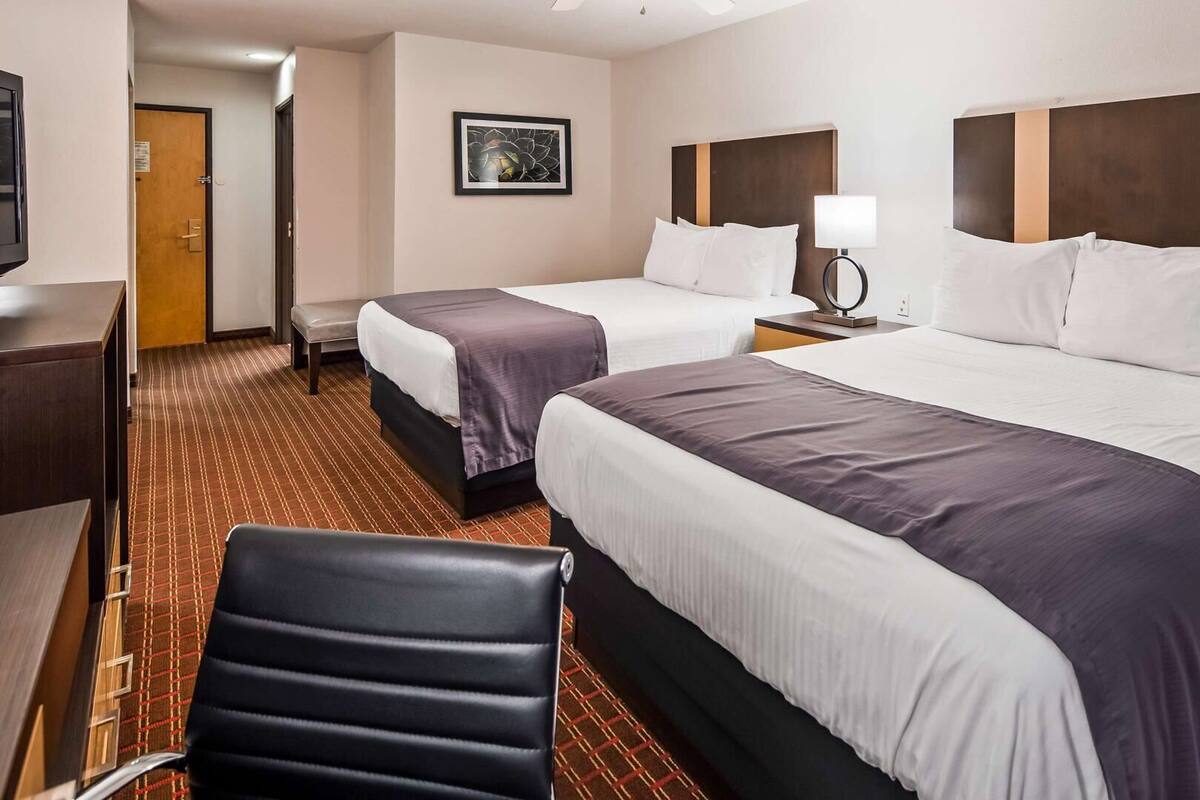 A hotel room with two beds, a desk, a chair, a nightstand with a lamp, and a framed picture on the wall.
