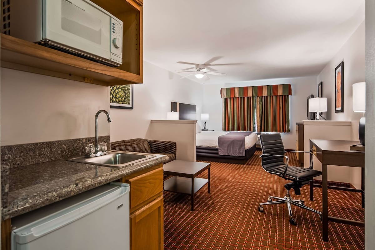 A hotel room with a kitchenette, a bed, a desk, a chair, and a ceiling fan.