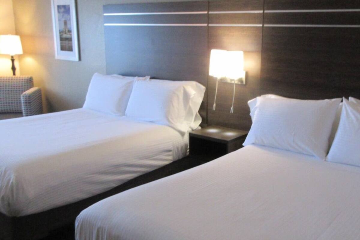 A hotel room with two beds, a nightstand, a chair, and wall-mounted lamps.