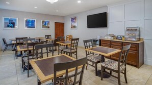 A dining area with multiple tables and chairs, a flat-screen TV mounted on the wall, and a small refrigerator with pastries. - Best Western Champions Forest Inn and Suites (Houston)