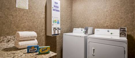 A laundry area with two Whirlpool washers, a wall-mounted soap and bleach dispenser, and a counter with folded towels and detergent boxes.