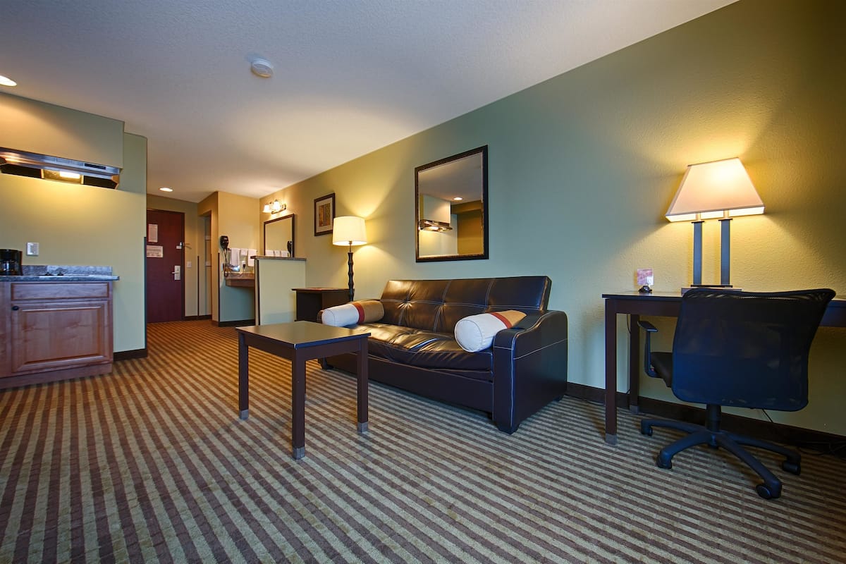 A hotel room with a sofa, a desk, a chair, a lamp, a mirror, and a kitchenette.