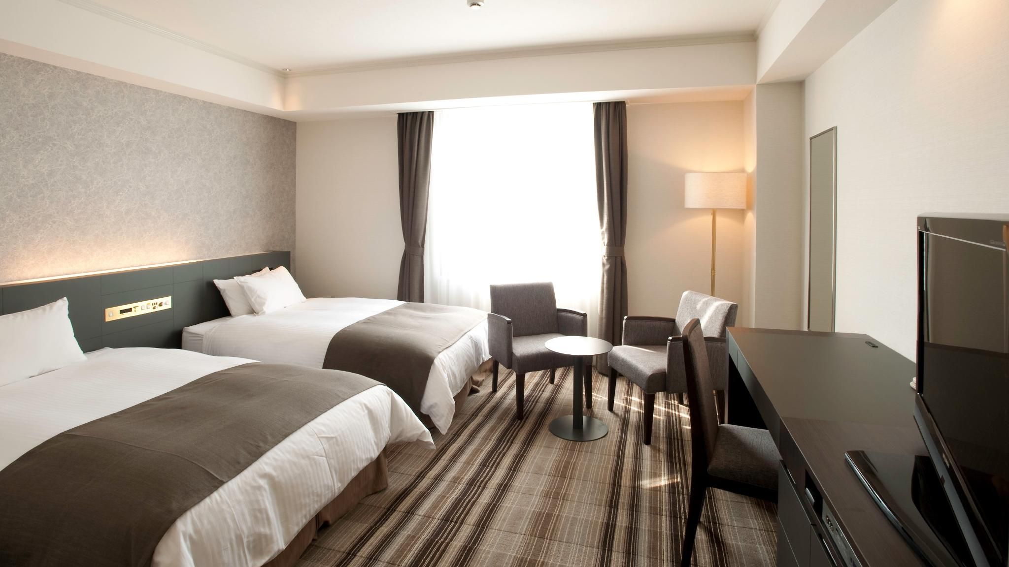 A hotel room with two beds, a desk, a chair, a TV, and a window with curtains.