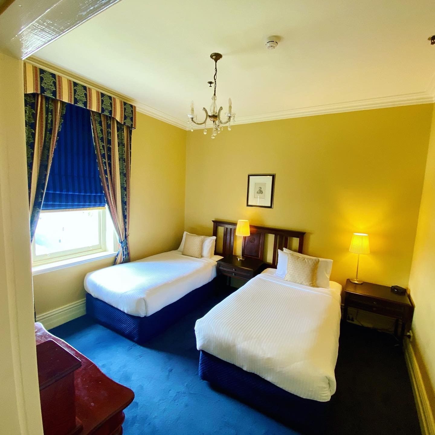 Habitación de hotel con dos camas, un candelabro, un cuadro en la pared y una ventana con cortinas.