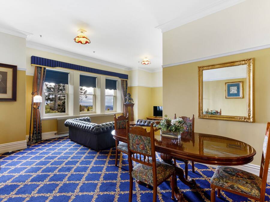 A room with a blue patterned carpet, a dining table with chairs, a large mirror, and a window with curtains.